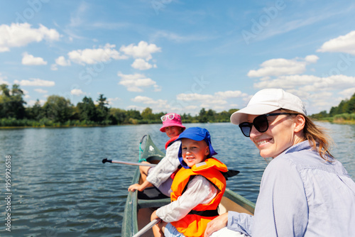 Woman two children enjoy having fun paddling canoe rent across lake river water wearing bright orange life vests hot sunny summer day. Healthy recreational activity lifestyle vacation trip journey