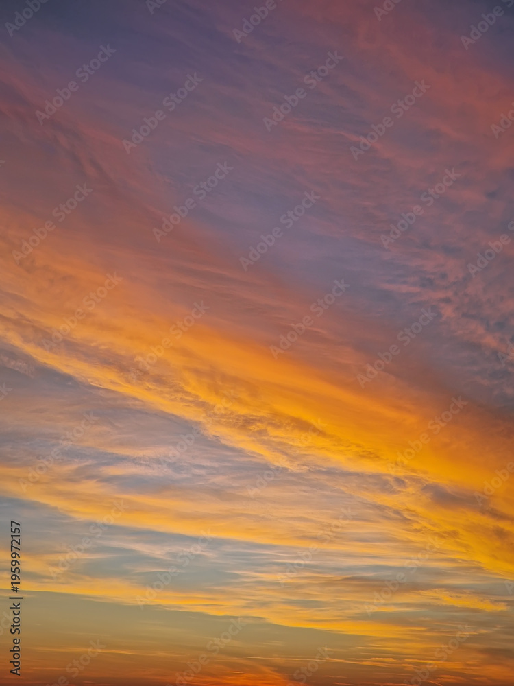 Fototapeta premium Vibrant sunset sky with multicolor layers of textured clouds. The horizon glows in fiery orange and yellow light, while the upper atmosphere transitions into soft, sweeping lavender and indigo tones