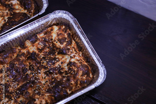 Close up shot of a golden brown crusty pie sprinkled with white sesame seeds. The savory pastry is presented in a silver foil container on a neutral background. 