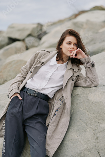 Young woman in textured coat and white shirt leans on rock outdoors, looking sideways with thoughtful expression. Casual fashion, nature, and relaxed mood in natural light.