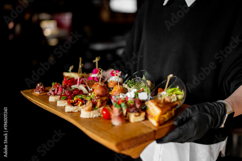 A waiter in black gloves holds a wooden tray of gourmet appetizers, including bruschetta, sliders, and salads in glasses. Elegant catering and finger food for an event or celebration