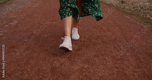 close-up of female legs wearing white sneakers walking on footpath. following legs camera from behind front side. Young beautiful woman walking in long green dress at summer nature. Ukrainian people