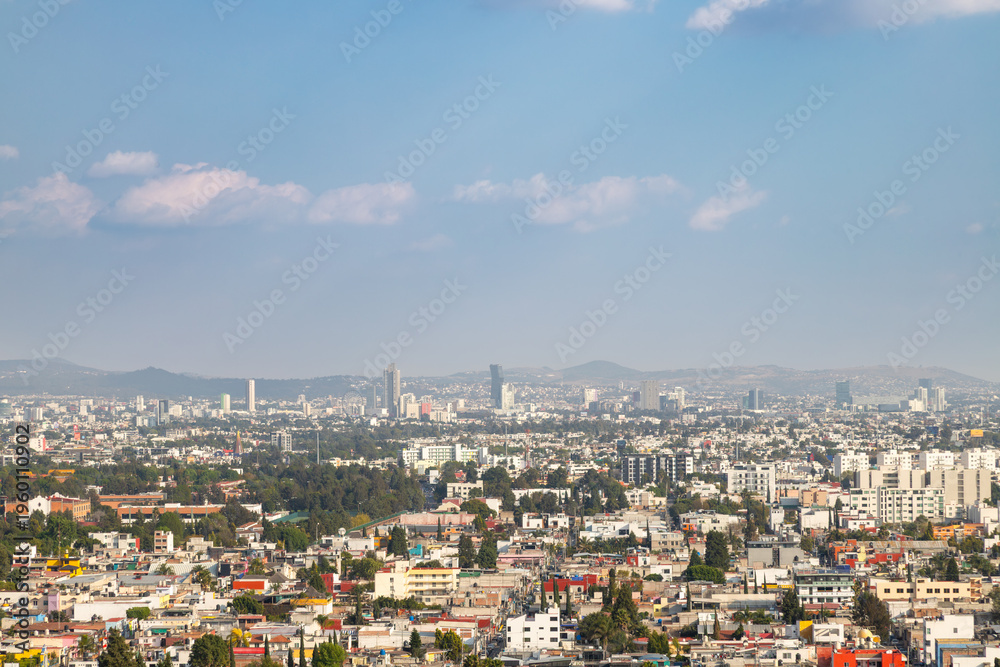 Obraz premium Dense urban panorama of Cholula with colorful residential buildings and distant Puebla skyline featuring skyscrapers and Ferris wheel under hazy blue sky in Cholula, Puebla, Mexico