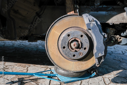 A car brake disc with visible rust and dust, mounted on a vehicle lifted by a blue jack. Brake caliper and mechanical components in focus, high performance car brake system condition checking