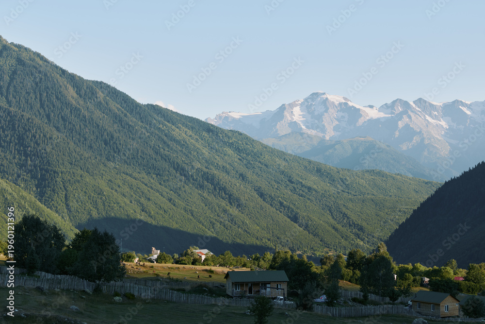 Obraz premium A tranquil mountain valley scene unfolds with green hills, distant snow capped peaks, and a clear blue sky. Meadows dot the landscape as rustic cottages sit beside stone walls and gentle sunlight