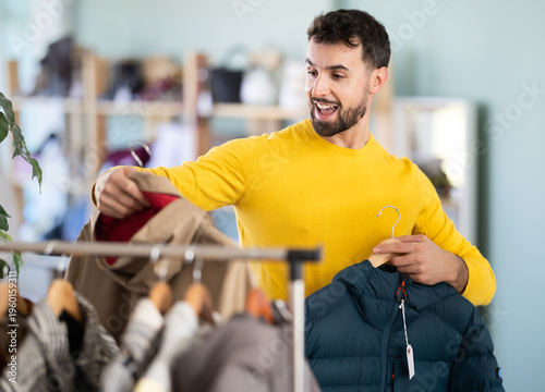 Quadro su tela Young man buyer choosing down jacket or raincoat in clothing store