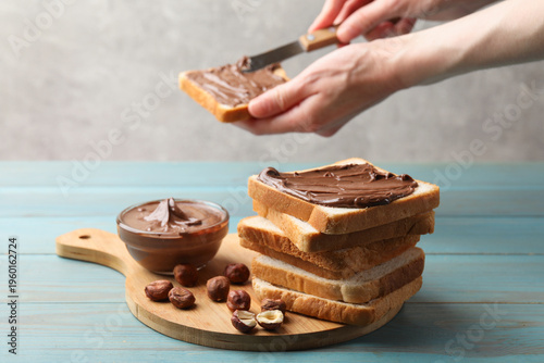 фотографія Woman spreading chocolate paste onto toast at light blue wooden table, selective