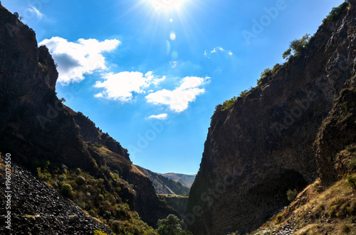 Deep, narrow canyon of Garni Gorge with steep, rocky walls under bright sun. Sky is blue with scattered white clouds, while the rugged slopes are dotted with sparse green vegetation and loose stone