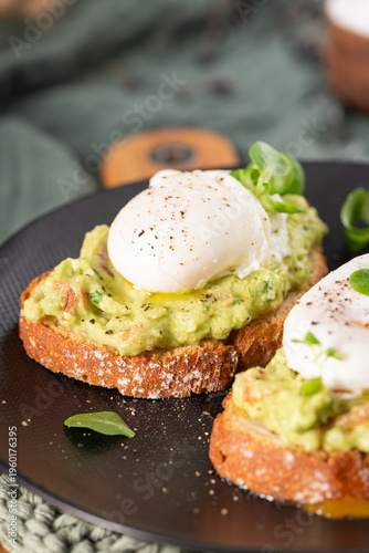 A vertical close up of a gourmet breakfast featuring crusty sourdough bread topped with creamy avocado mash and a soft poached egg with black pepper and fresh microgreens on a black plate