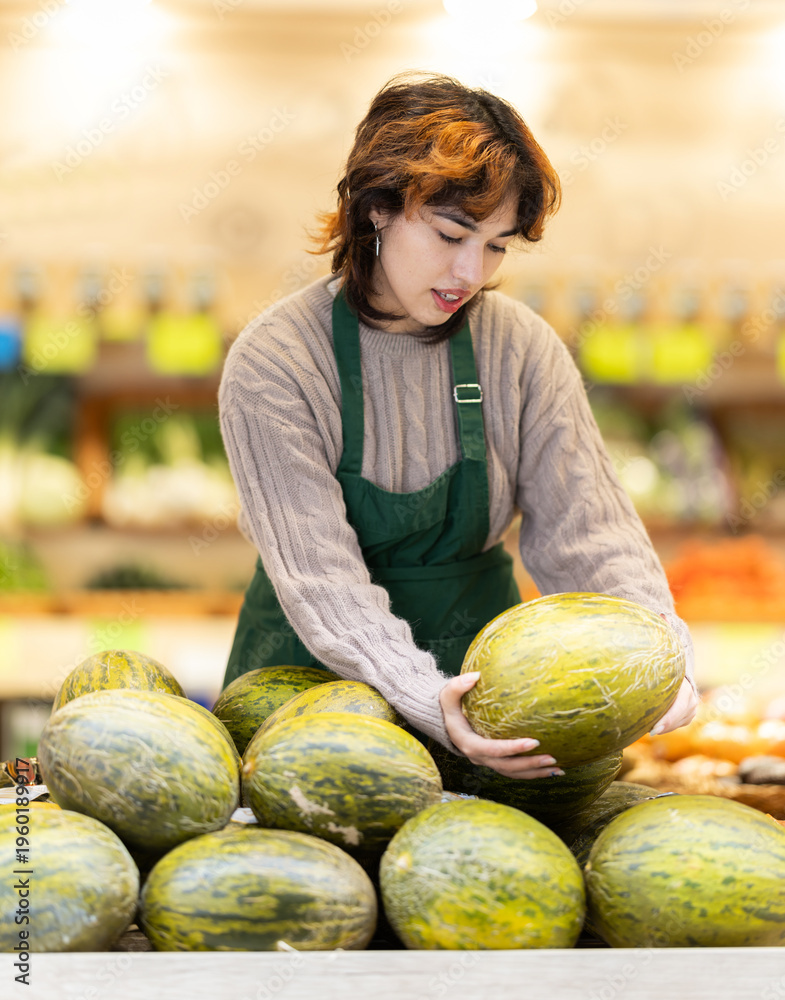 Fototapeta premium Seller in grocery vegetable shop arrange melon. Girl creates stock of fruit in display, forms showcase. Vegetable shop near house. Organic vegetable shop offer assortment, business case
