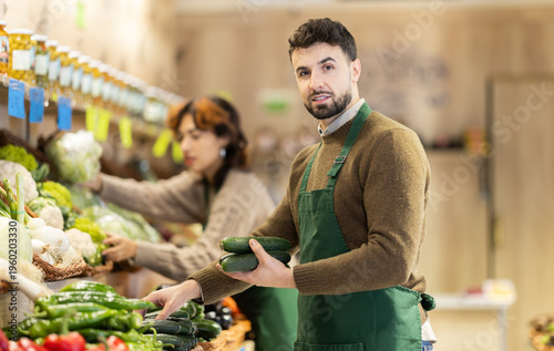 Fotografía Attentive young male seller putting cucumbers on stand in fruit and vegetable ma