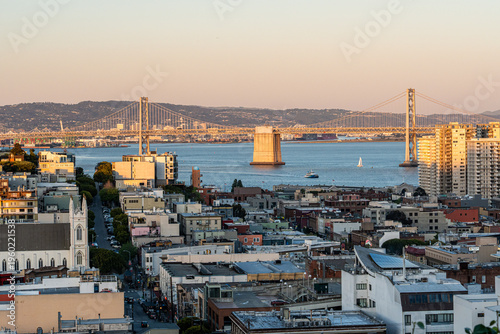 The San Francisco-Oakland Bay Bridge gleams in the warm sunset light above the historic rooftops and church spires of the North Beach district