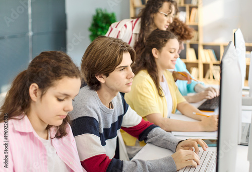 Student teacher teenagers in classroom with computers. Focused students learn coding together as the instructor guides teamwork and provides training. Clear concept of digital education.