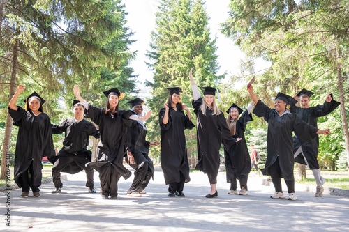 Happy multinational young people in graduation gowns jumping for joy outdoors. 