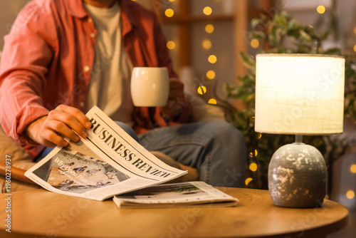 Young man with cup of tea taking newspaper from table at home in evening, clo...