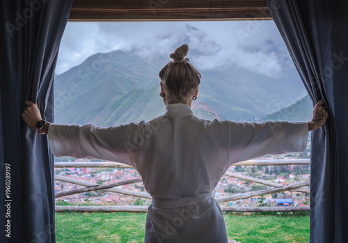 Woman tourist wearing a white bathrobe looking through a window at the Sacred Valley of the Incas in Peru. Travel and relaxation concept in a luxury accommodation, representing tourism, wellness.