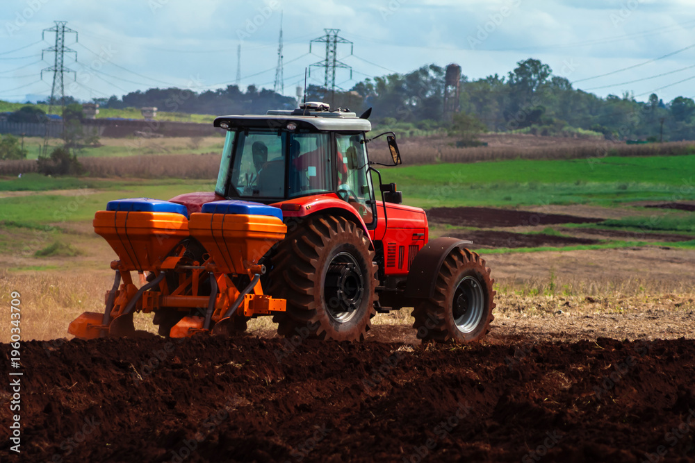 Fototapeta premium Red tractor with a seeder plowing a field in a rural landscape, with power transmission towers in the background, symbolizing modern agriculture and energy infrastructure. 