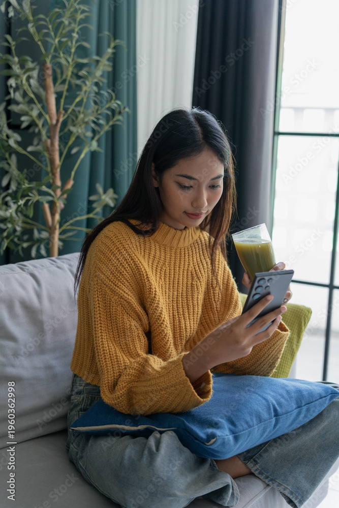Obraz premium Asian woman in sweater using smartphone and drinking green vegeteble juice