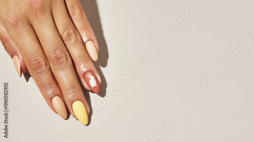 A close-up of a woman's hand with long, almond-shaped nails. The nails are painted in pastel colors, including yellow and a nude shade. The background is neutral.