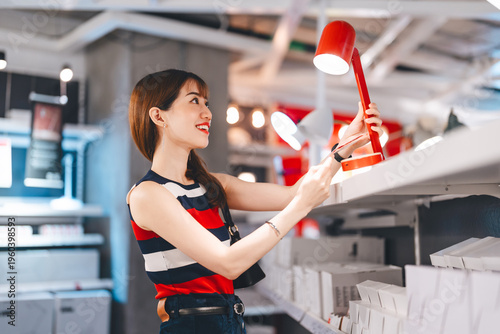 Adult asian woman choosing lamp light product home interior shopping buying new furniture at warehouse superstore
