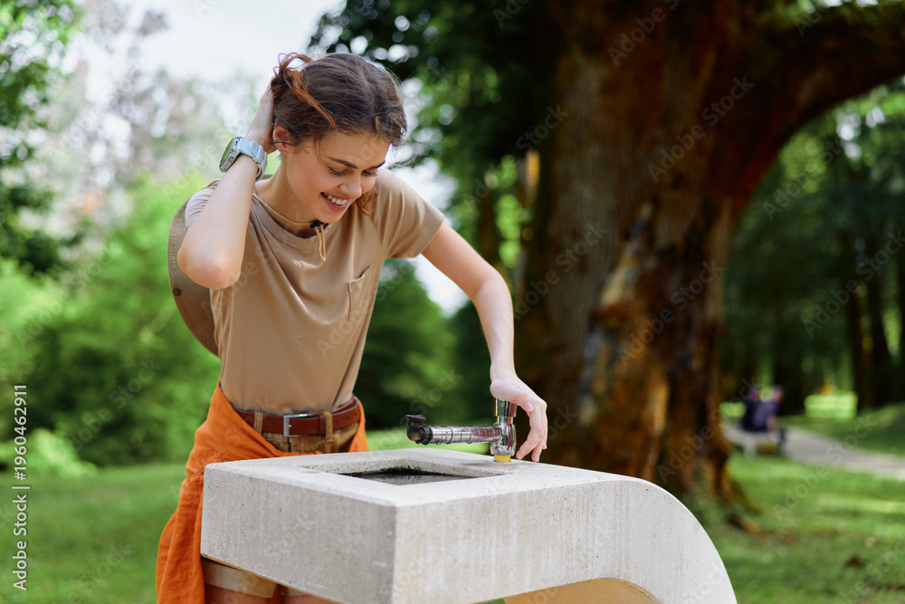 Obraz premium Young woman smiling and drinking from a public water fountain in a green park, casual summer outfit, pressing the spout for hydration, outdoor leisure and healthy lifestyle moment.
