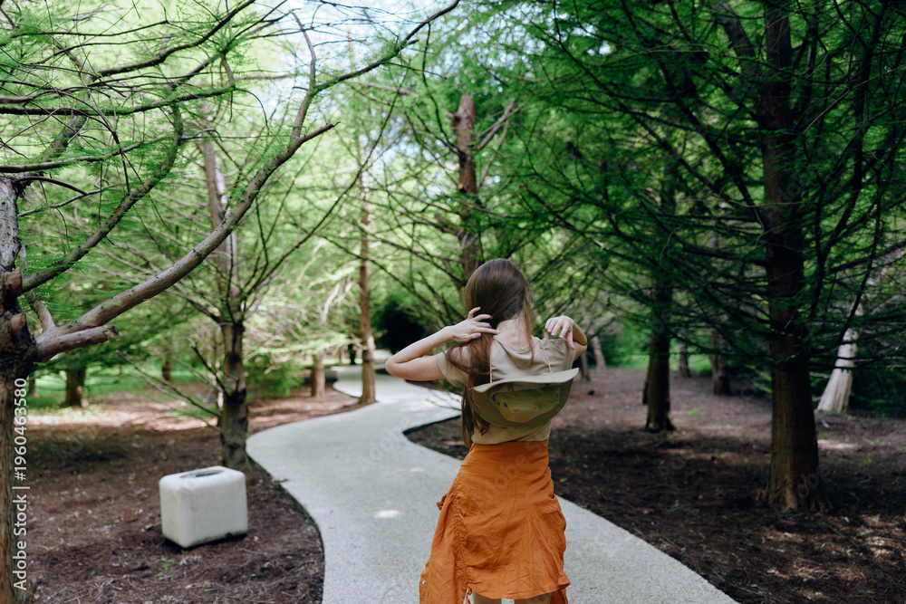 Naklejka premium Woman walking on winding path through forest trees, back view with hat and orange skirt, outdoors scene of summer park trail, scenic nature stroll and relaxed leisure.