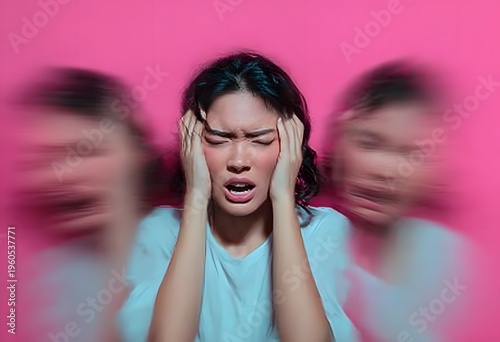 A young woman suffering severe mental anguish or intense headache gripping her head on a vivid pink background with blurred side figures