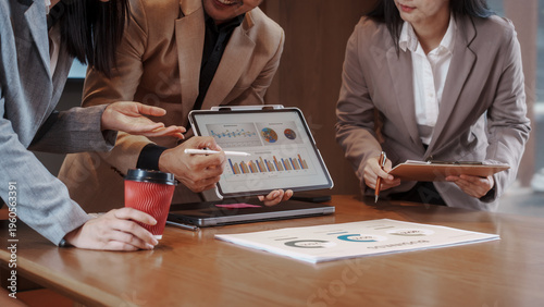 Smiling business team collaborating over documents in modern office with data charts on screen, discussing strategy and analytics during meeting.