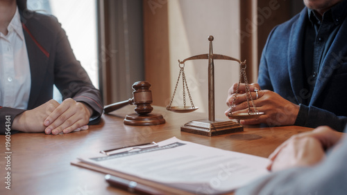Close-up of legal consultation meeting with attorneys at desk, scales of justice and gavel in focus, paperwork in foreground, warm office light.