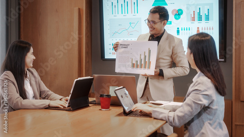 Billede på lærred Business team meeting in modern conference room, presenter shows sales chart to colleagues with laptops and tablet, large screen with analytics dashboards in background
