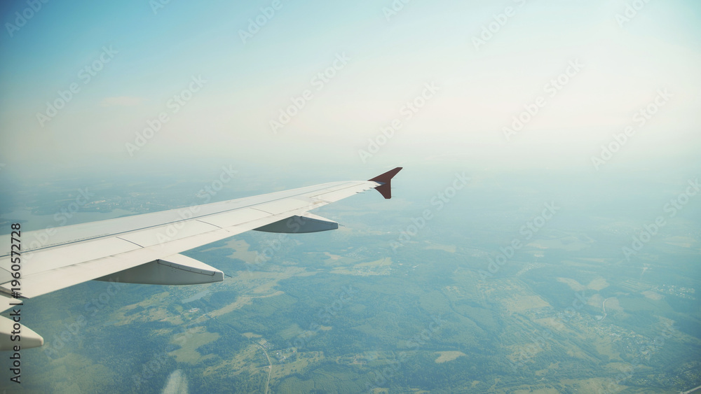 Fototapeta premium Airplane wing hovering over verdant countryside, revealing patchwork fields, rustic villages, and serpentine roads during sunny aerial journey