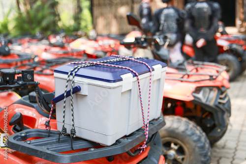 White cooler secured with strap on front rack of an orange all terrain vehicle stands ready for an exciting outdoor adventure alongside parked fleet of quads with riders