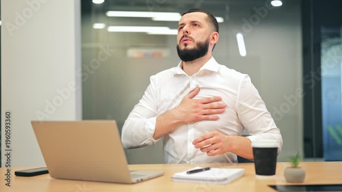 Professional at desk feels discomfort, placing hand on chest. Suggests medical concern, pain, or intense stress while working on laptop in modern office environment.
