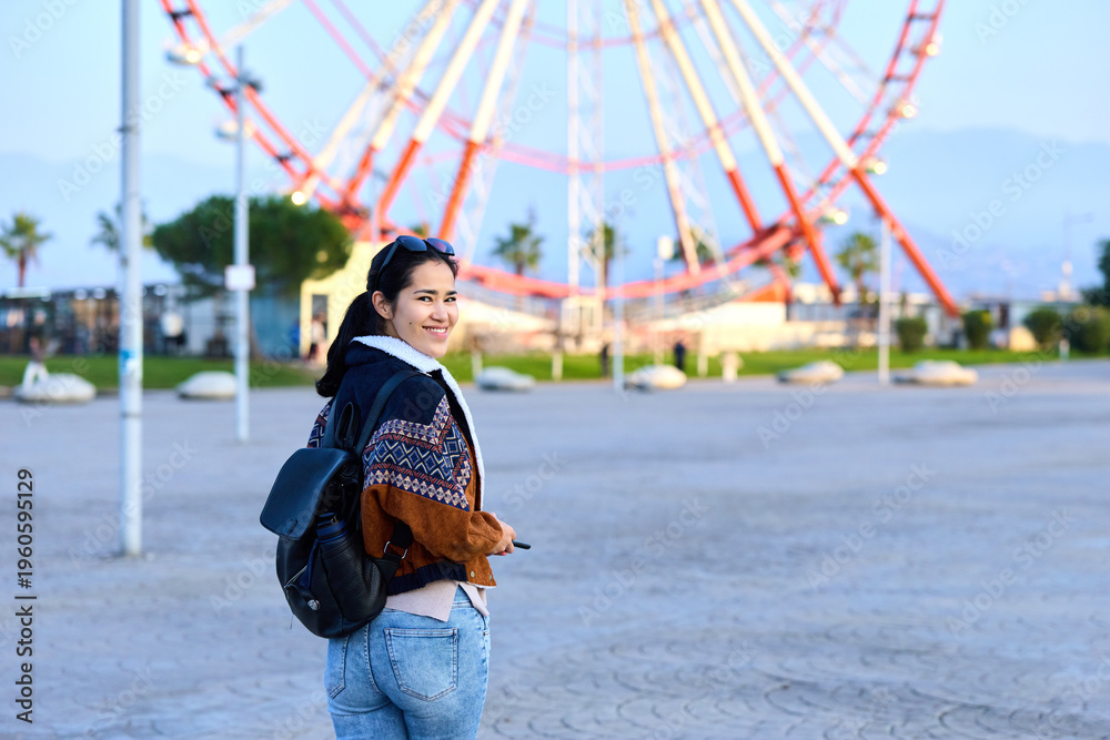 Fototapeta premium Girl backpack amusement park roller coaster outdoor casual youth smiling as she strolls near the ride, capturing a bright, airy moment of exploration and leisure in a sunny outdoor setting