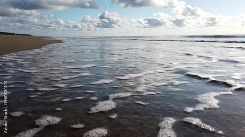 Beach shoreline with gentle waves lapping against wet sand, reflecting clouds and blue sky, creating a serene coastal landscape in a natural setting