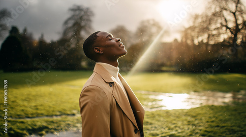Young man in beige trench coat stands in spring rain, face raised to sky, eyes closed, smiling peacefully, warm golden sunlight breaking through dramatic storm clouds over lush green park.