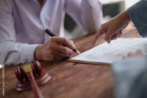 Fotografie Person's hand signing a legal document on a wooden table, with another person po