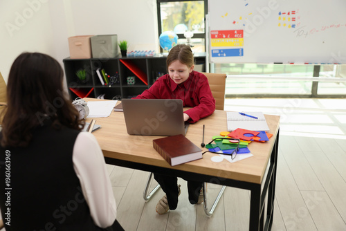 A child in a red shirt focuses on a laptop during an online learning session. A teacher observes from a nearby chair, surrounded by colorful classroom materials and bright decorations.