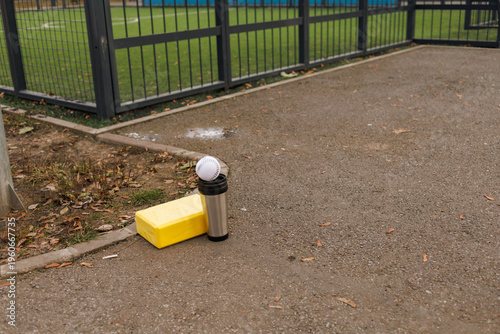 A white ball rests on top of a small black container beside a yellow box near a sports field