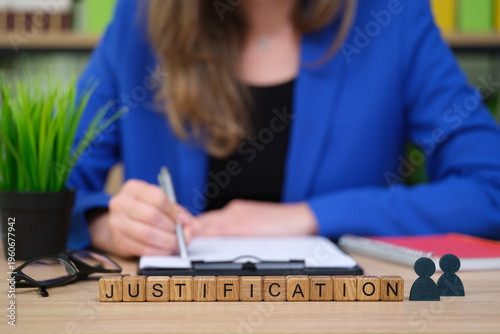 In a cozy setting, a person in a blue blazer writes on a clipboard. Wooden blocks neatly display the word 'justification.' Nearby, a plant adds a touch of nature and calm.