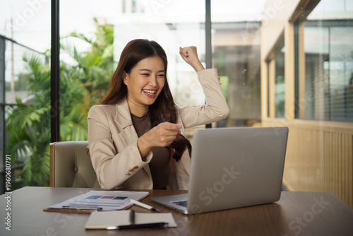 Happy professional businesswoman raising fists in triumph after good news on computer. Business success and goal accomplishment theme.