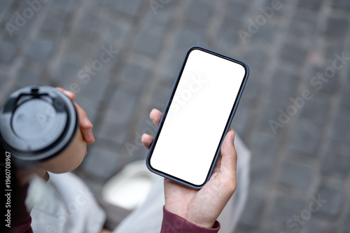 Top view of a man's hand holding white screen phone with coffee while sitting on bench over sidewalk