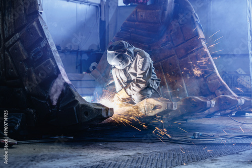 Slika na platnu Welder kneeling cutting large excavator bucket inside factory bay, recovery work