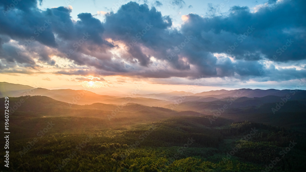 Obraz premium Aerial view breathtaking sunset over forest. Sun's rays break through dramatic clouds, casting golden glow across rolling hills and valleys. Distant mountains creating serene and picturesque scene.