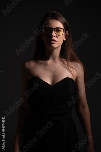 Young woman with glasses wearing a black strapless dress in low light, standing against a dark background, portraying elegance, mystery, and a confident mood in a fashion studio portrait