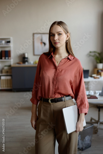 Portrait of young adult businesswoman holding laptop and standing in office, presenting professional confidence and workplace readiness. Ideal for career growth, recruitment, corporate branding, verti