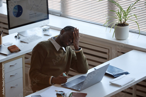 Black young adult businessman holding head while working on laptop at office desk, showing stress and fatigue. Useful for burnout, workload pressure, mental health, corporate wellness themes