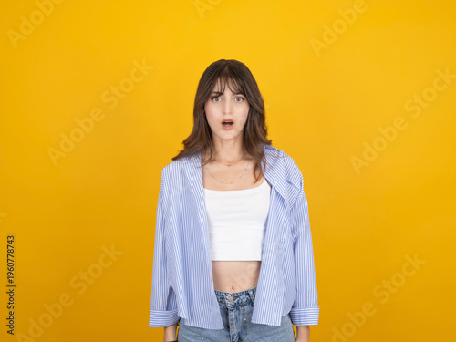 Tableau sur toile Surprised young woman with shocked expression looking forward, wearing striped shirt and denim, standing against bright yellow studio background with copy space