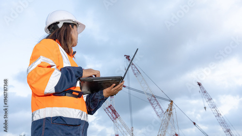 A worker stands at a construction site looking at a laptop while cranes are active in the background. The worker wears safety gear and focuses on the task