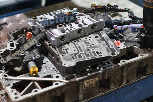 Detailed view of a disassembled automatic transmission valve body with complex metal parts and electrical wiring on a workbench in a professional mechanic workshop.